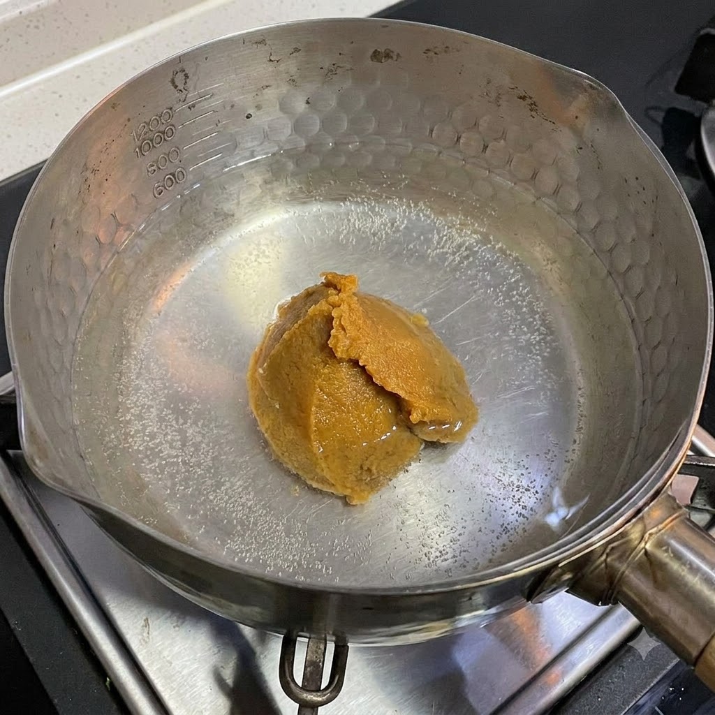Une cuillerée de pâte de miso blanc en train de se dissoudre dans une casserole deau chaude sur une cuisinière.