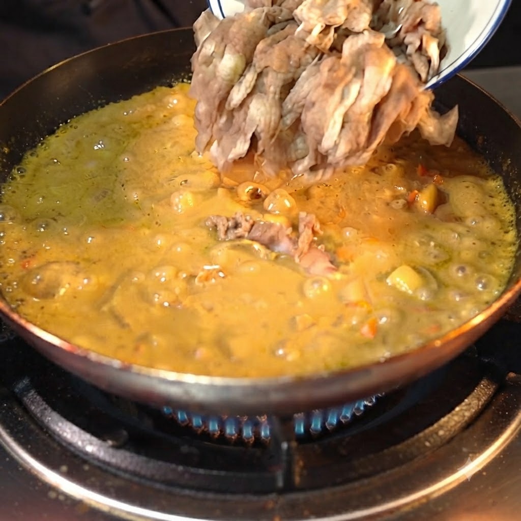 Des tranches de bœuf pré-transformées versées dun bol blanc dans une sauce au curry jaune frémissante et épaissie dans une poêle sombre.
