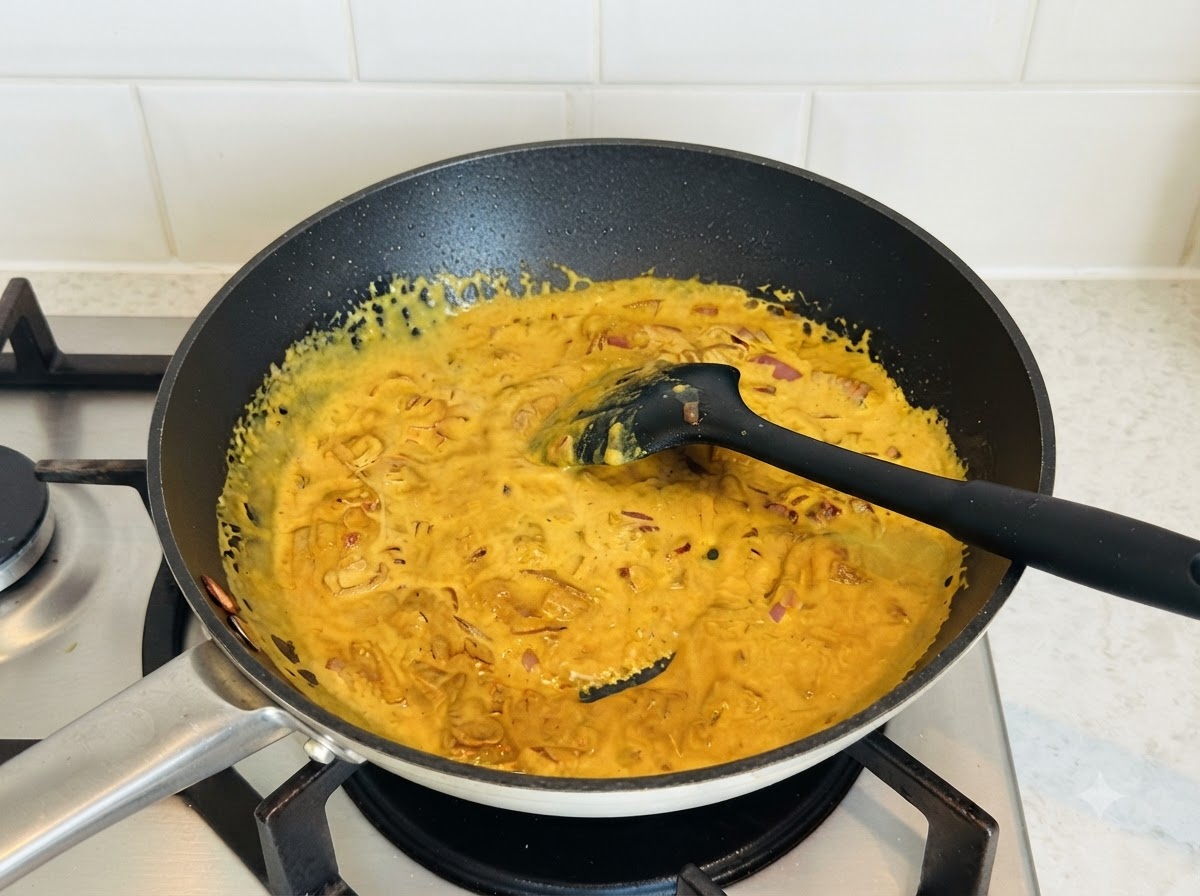 Une sauce au curry jaune et riche en cours de remuage dans une poêle foncée avec une spatule en silicone noire.