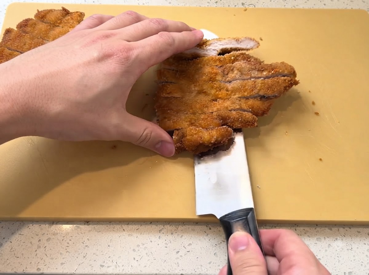Une côtelette de porc frite dorée en train dêtre tranchée en lanières avec un couteau bien aiguisé sur une planche à découper jaune.