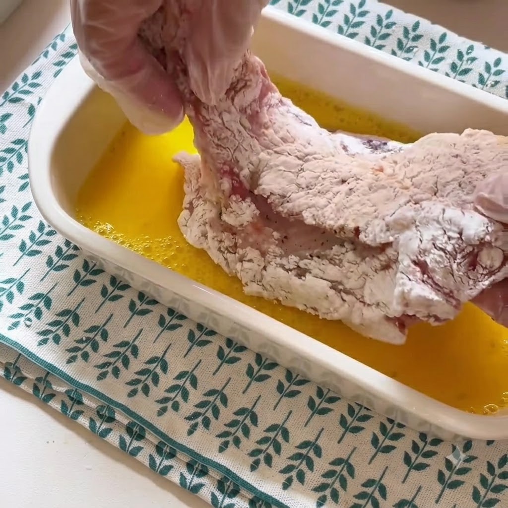 Une main gantée soulevant une côte de porc enrobée de fécule hors dun récipient contenant du liquide dœuf battu jaune.