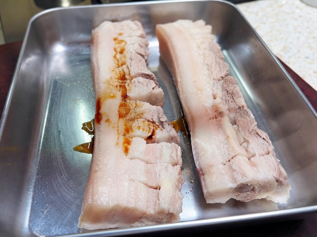 Deux morceaux de poitrine de porc quadrillés sur un plateau en métal avec de la sauce soja foncée versée sur la viande.