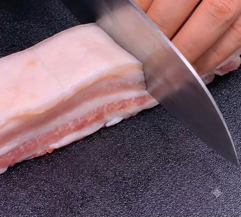 Un couteau bien aiguisé faisant des coupes précises et régulières dans une lanière de poitrine de porc sur une planche à découper sombre.