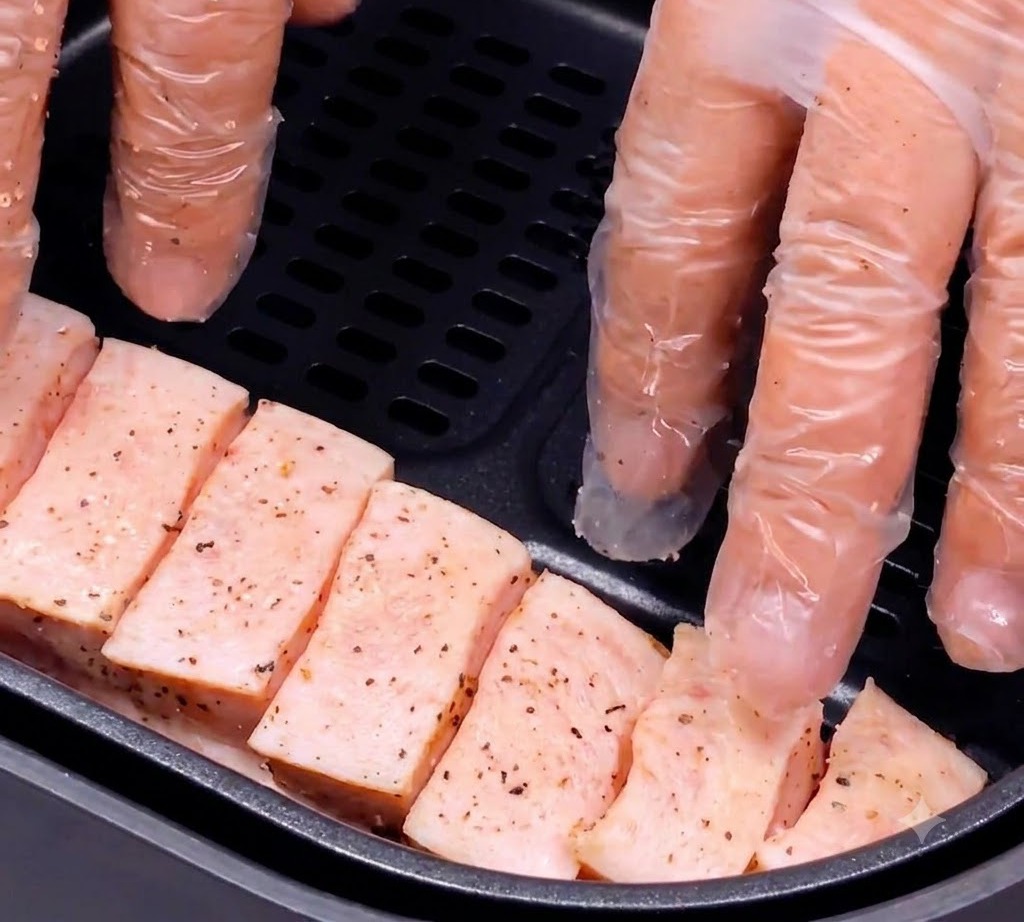 Lanières de poitrine de porc assaisonnées placées avec précaution dans le panier sombre dun air fryer par des mains gantées.