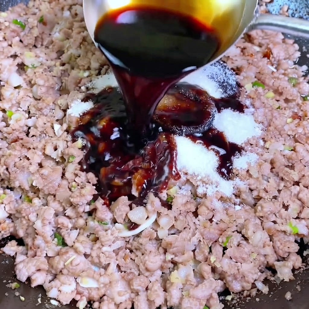 De la sauce soja foncée versée dune louche argentée dans un wok contenant du porc haché et des assaisonnements blancs.