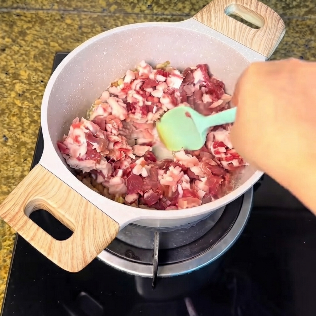 De la viande de porc en dés versée dans la casserole de graisse fondue et mélangée avec une spatule verte.