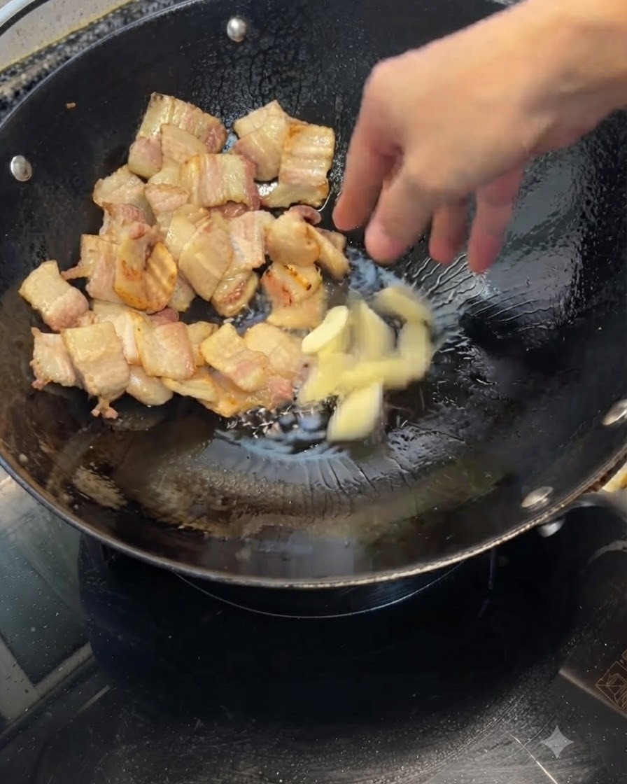 Gousses dail écrasées jetées dans un wok contenant des morceaux de poitrine de porc dorés.