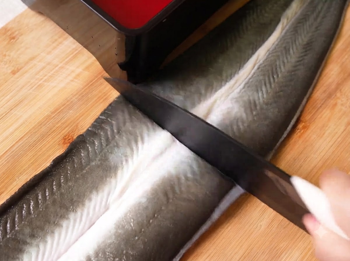 Un couteau tranchant découpant une grande anguille crue ouverte en papillon en trois sections sur une planche à découper en bois.