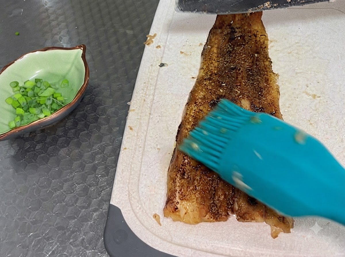 Filet danguille cuit coupé sur une planche blanche à côté dun petit bol contenant de la ciboule hachée.