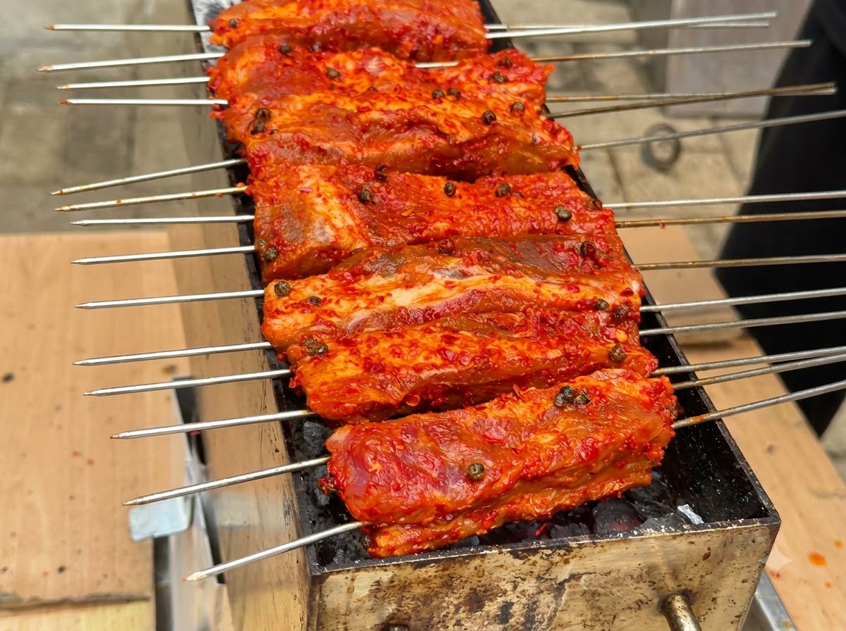 Plusieurs brochettes de côtes enrobées de rouge alignées et grillant sur un foyer au charbon.