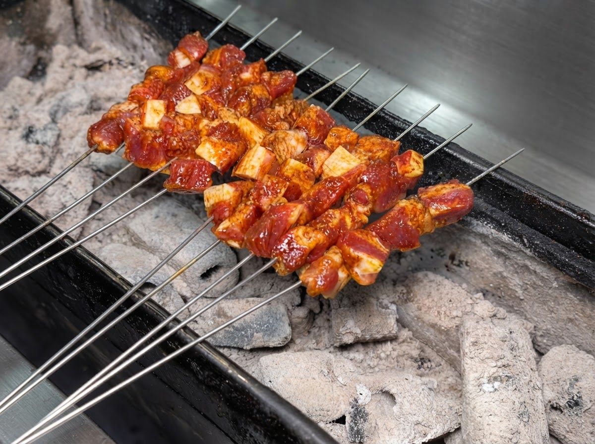 Plusieurs brochettes en métal de viande de porc assaisonnée disposées proprement sur un gril à charbon ardent.