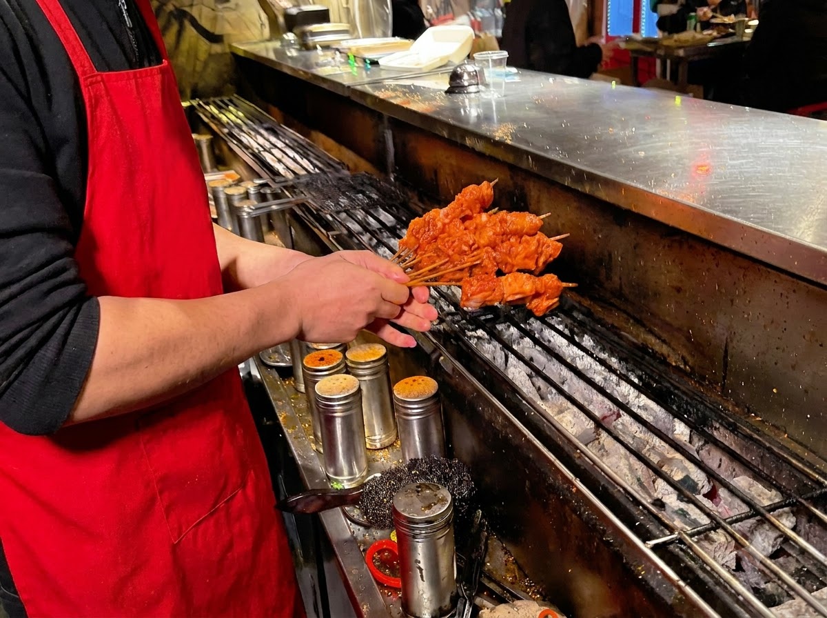 Personne portant un tablier rouge plaçant des brochettes de porc sur un gril à charbon allumé.