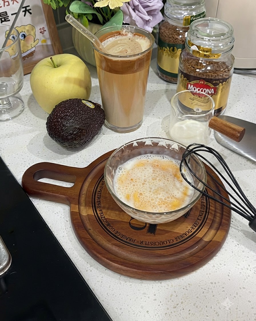 Un bol en verre rempli dun mélange mousseux battu dœuf et de lait à côté dun fouet noir.