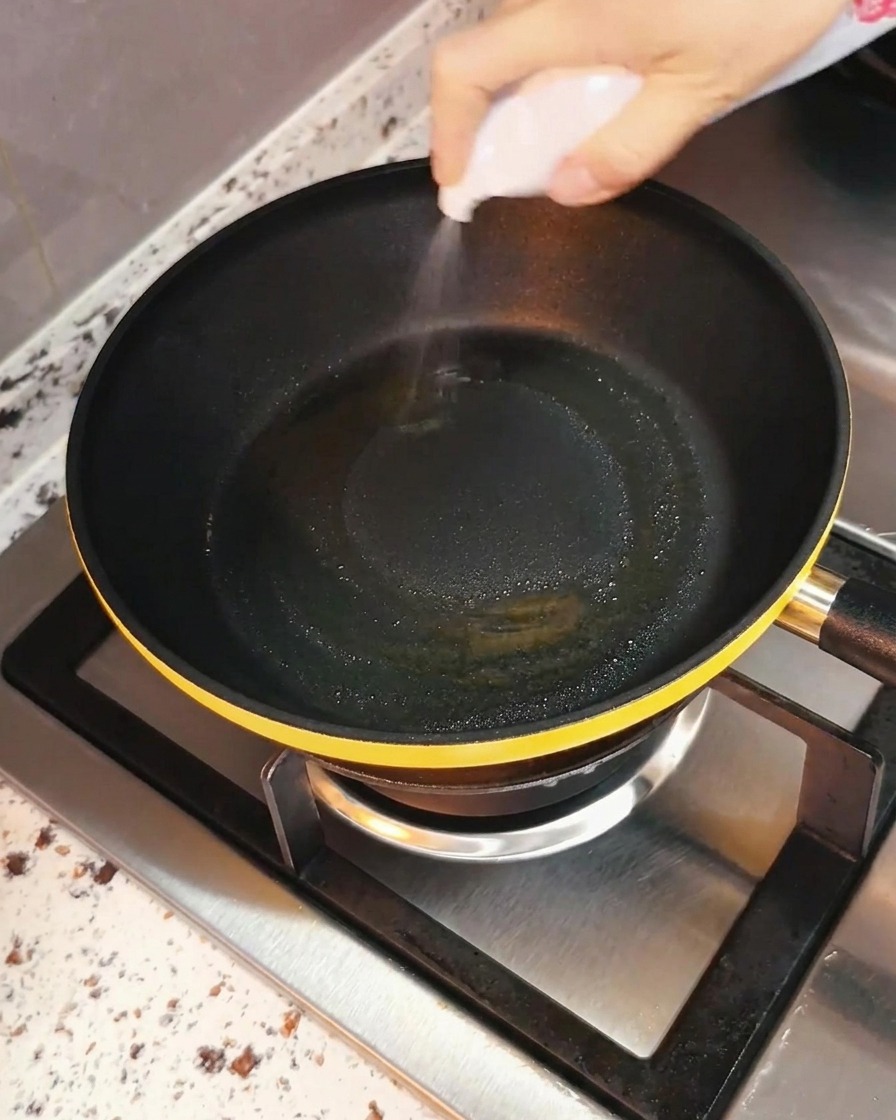 Une main vaporisant uniformément de lhuile de cuisson dans une poêle noire posée sur un brûleur de cuisinière.