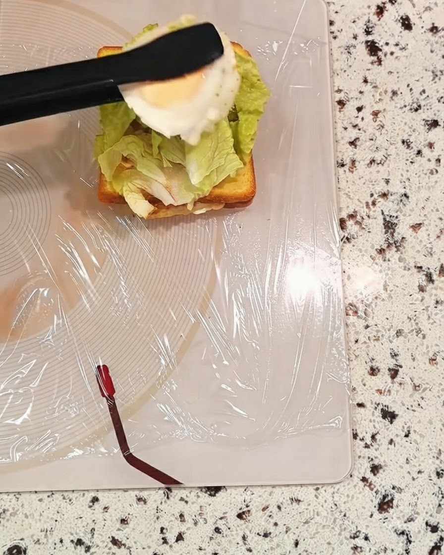 Une paire de pinces noires tenant un œuf cuit au-dessus dune couche de sandwich avec de la laitue.