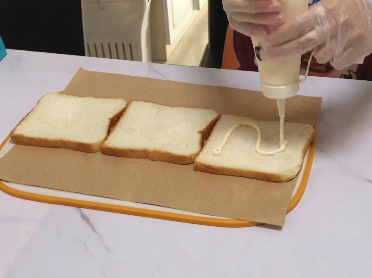 Assaisonnement à la sauce blanche versé en ligne ondulée sur une tranche de pain de sandwich posée sur du papier sulfurisé.