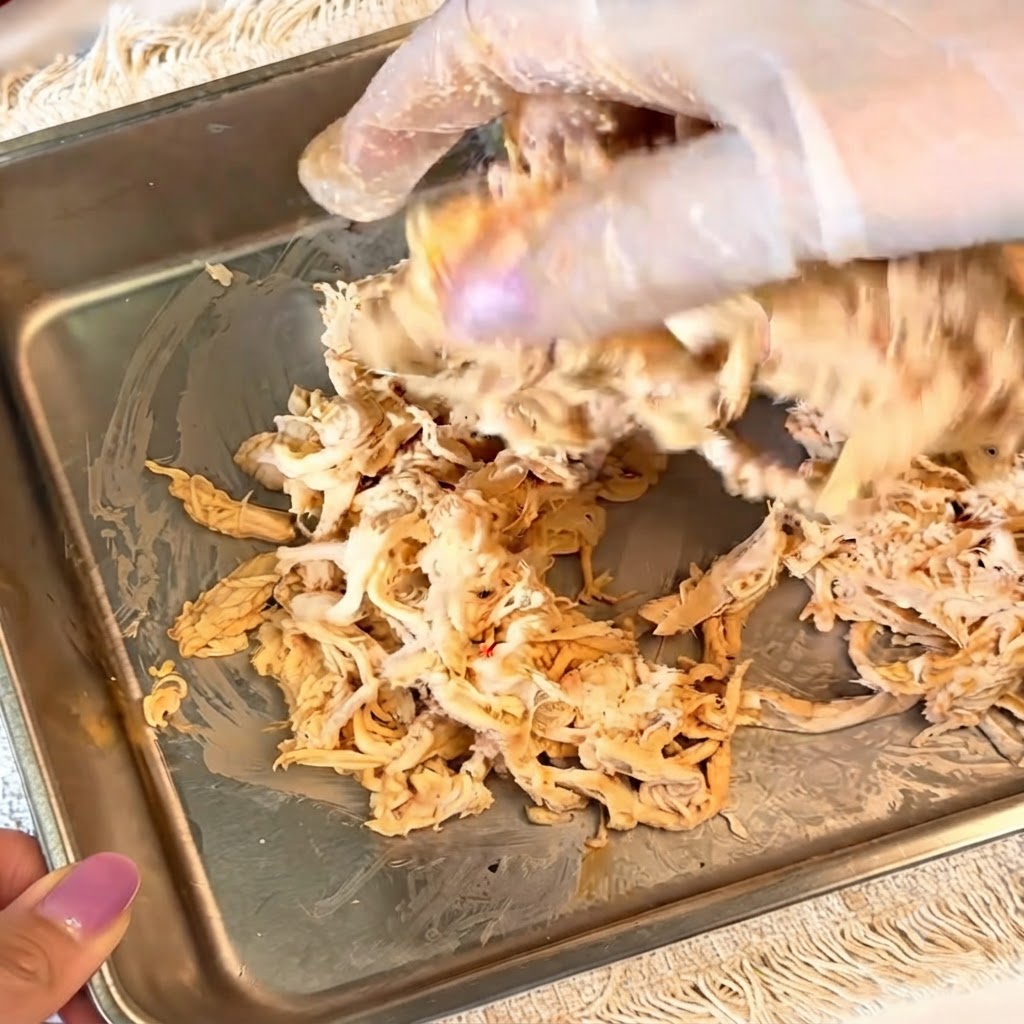 Des mains gantées mélangeant du poulet effiloché assaisonné dans un récipient métallique.