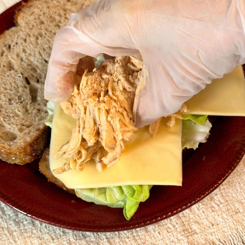 Des gants plaçant une portion de poulet effiloché assaisonné sur la couche de fromage du sandwich.