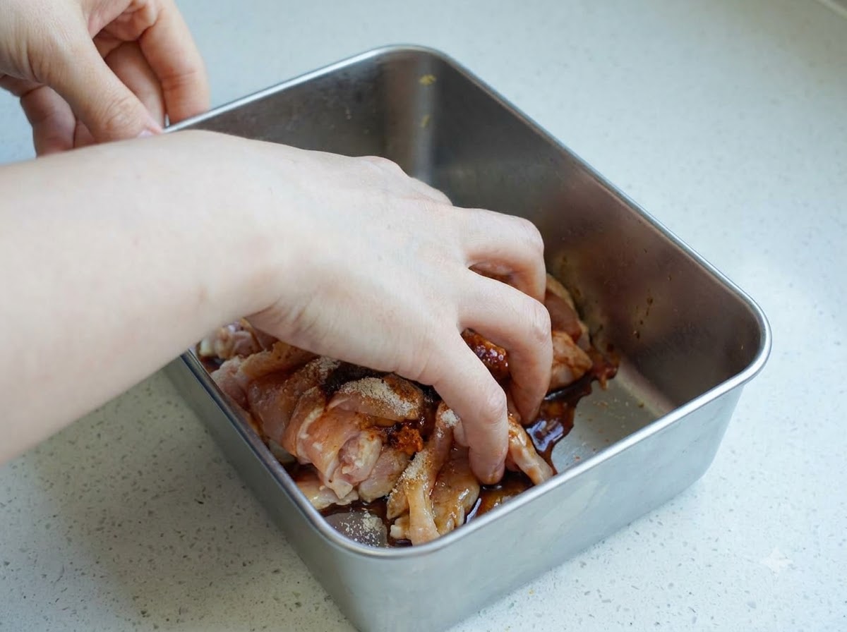 Une main mélangeant des morceaux de poulet cru assaisonnés à lintérieur dun récipient en métal.