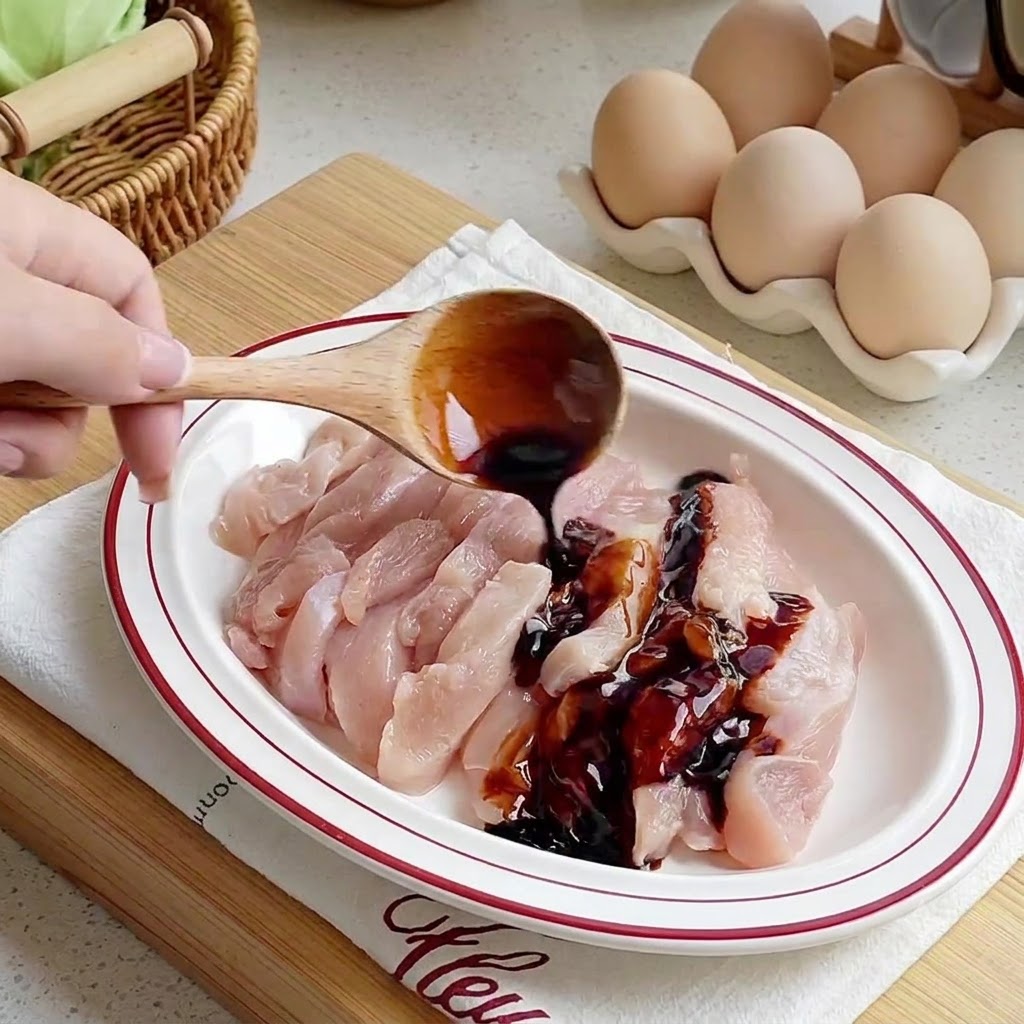 Des sauces versées à laide dune cuillère en bois sur des lamelles de poulet cru disposées dans une assiette blanche.