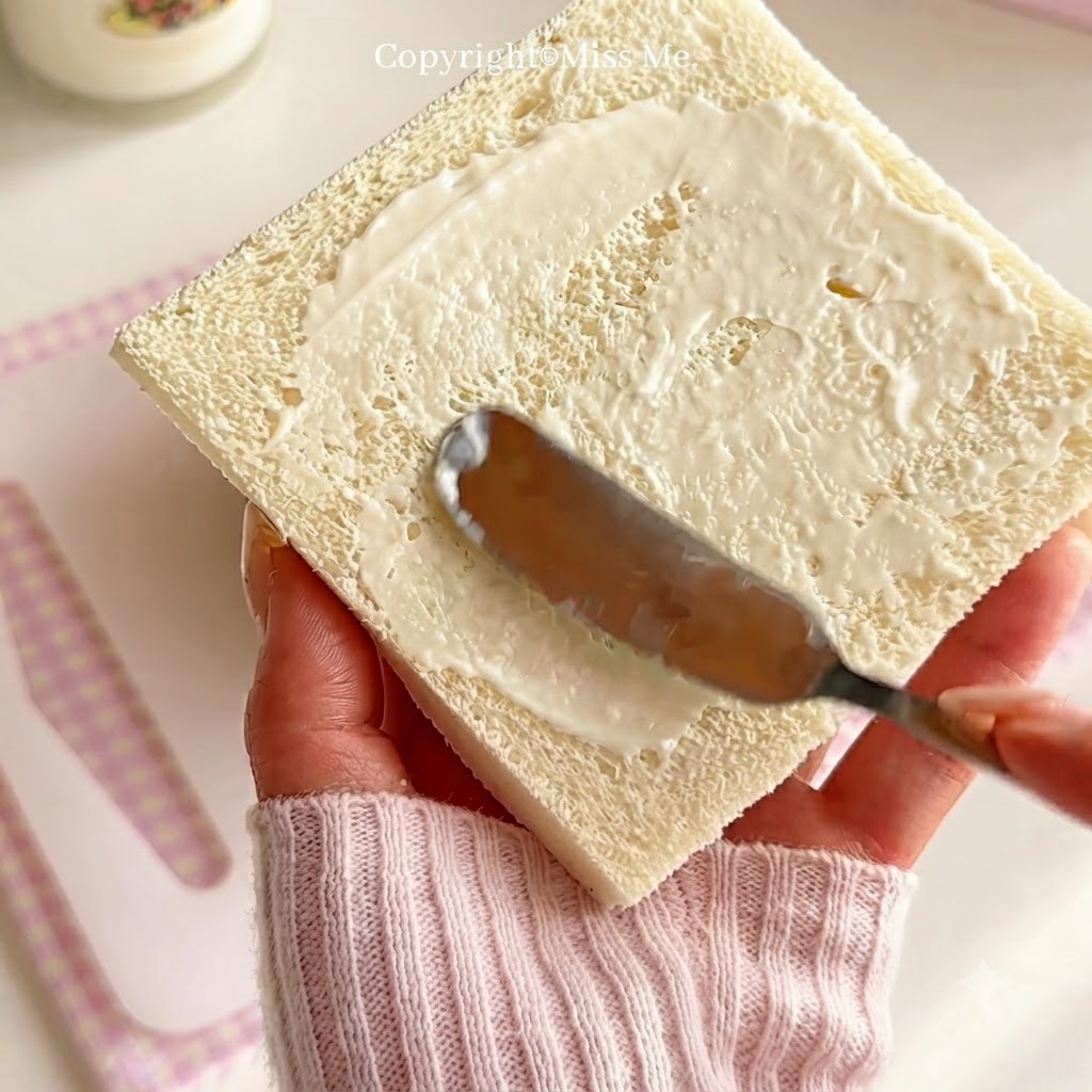 Une main tenant un couteau pour étaler de la sauce blanche uniformément sur une tranche carrée de pain de mie.