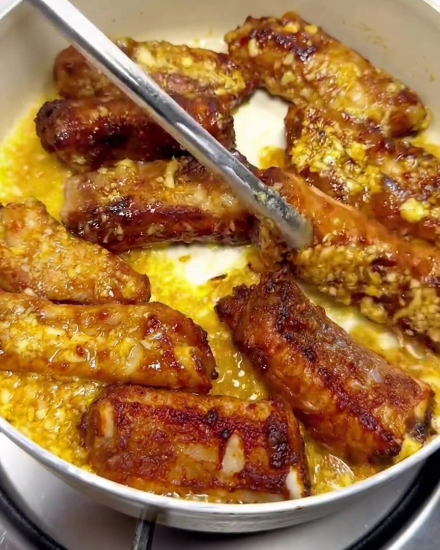 Des travers de porc retournés avec des pinces en métal dans une poêle, enrobés de sauce au beurre à lail.