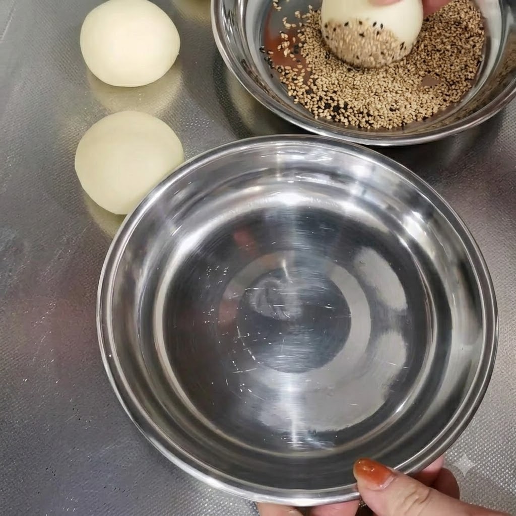 Une boule de pâte enrobée de graines de sésame dans un bol en métal à côté dautres boules de pâte préparées.