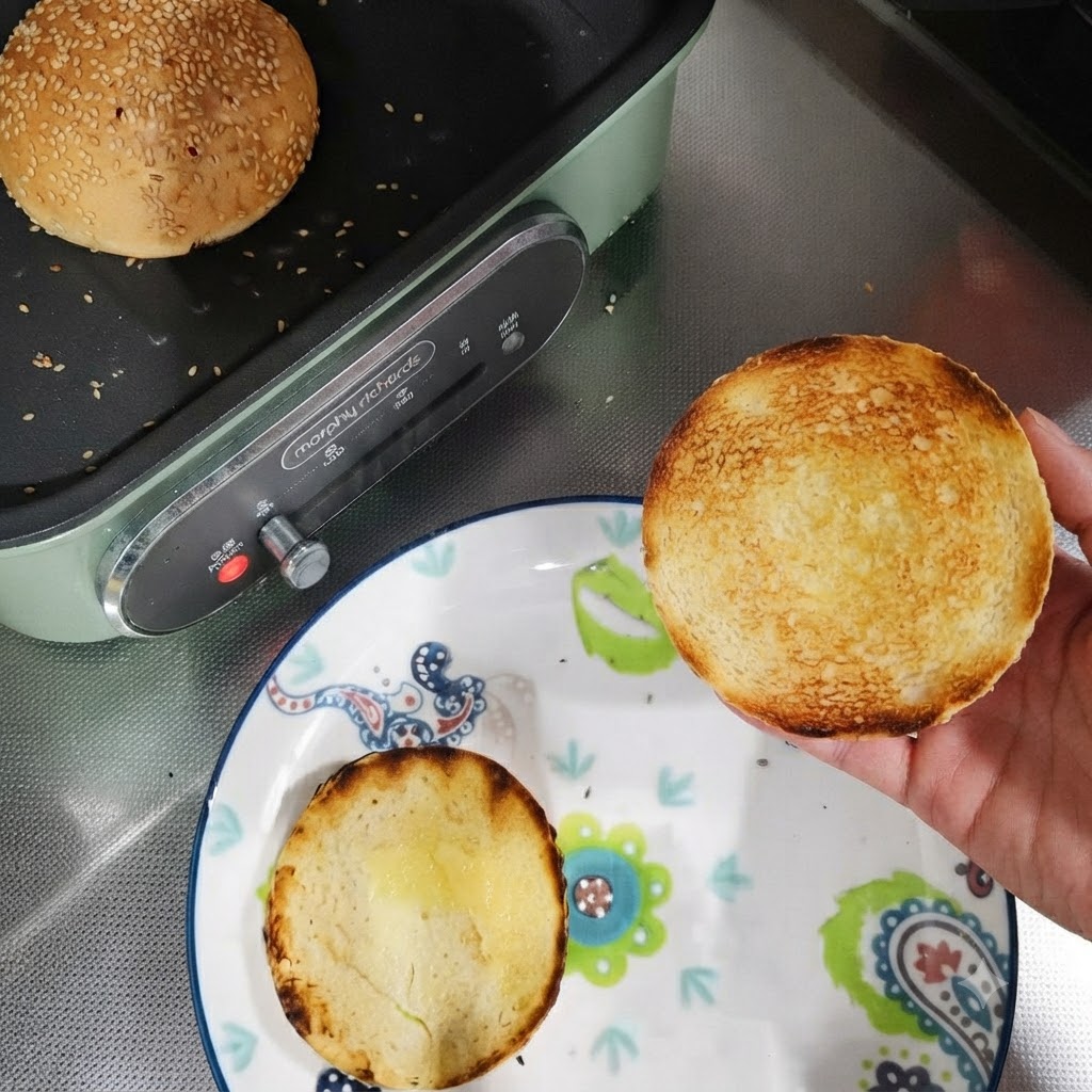 Moitiés de pain à burger toastées dans une assiette à côté dune poêle avec un pain entier en train dêtre toasté.