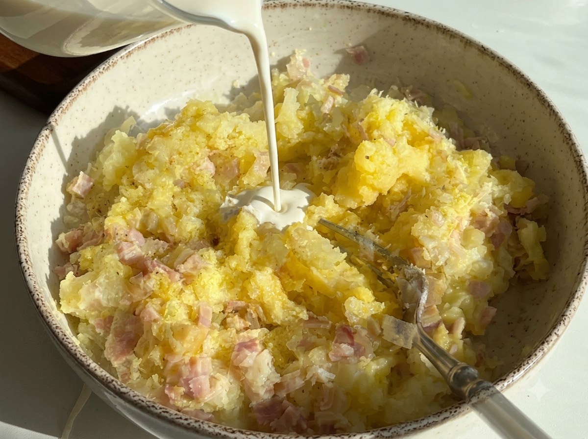 Crème fraîche versée dans un bol contenant de la purée de pommes de terre et des dés de jambon.