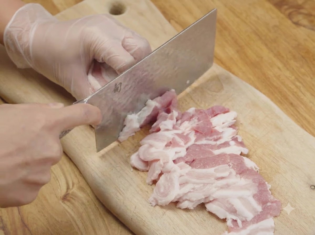De la poitrine de porc crue découpée en fines lanières sur une planche à découper en bois avec un couperet de cuisine.