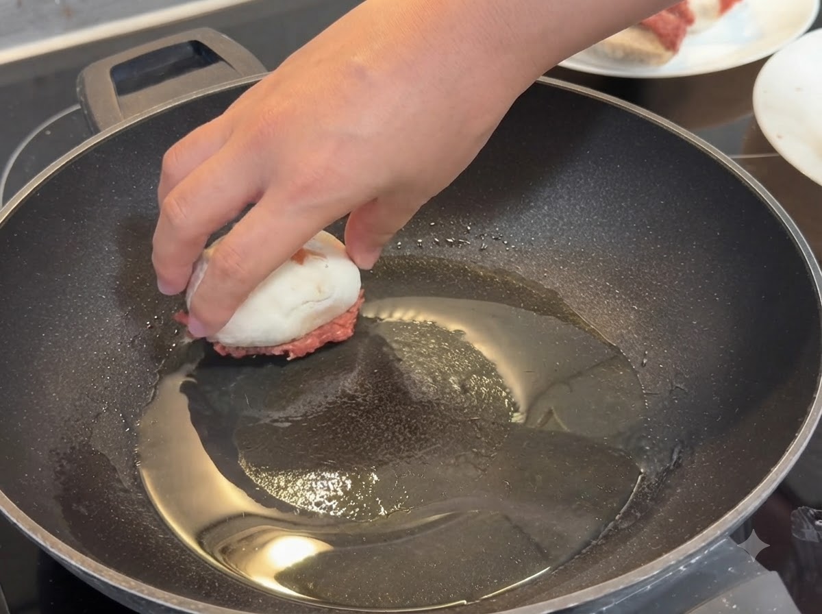 Une main plaçant un champignon farci côté viande vers le bas dans une poêle chaude huilée.