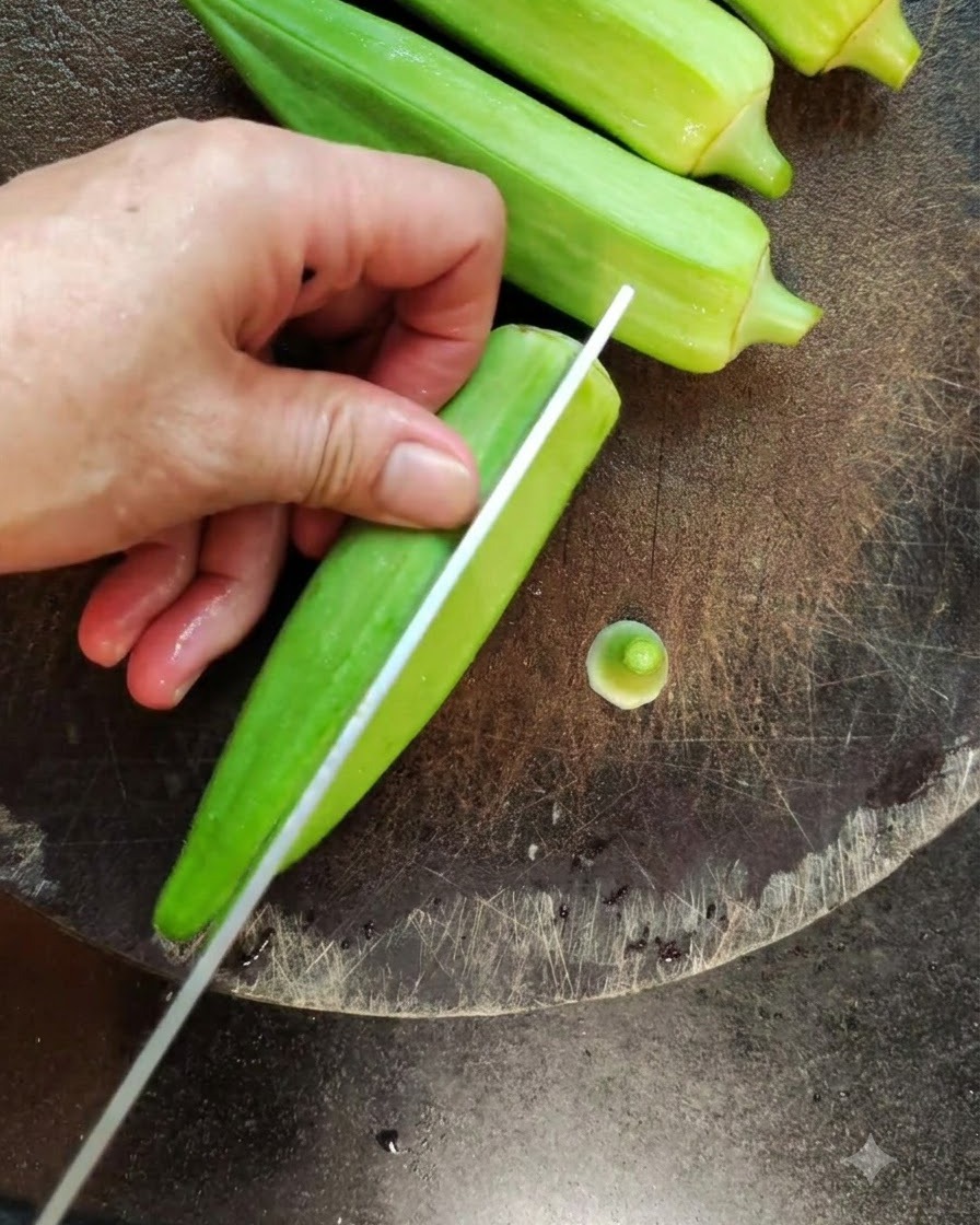 Gros plan dun couteau coupant un gombo vert frais en deux dans le sens de la longueur sur une planche à découper sombre.