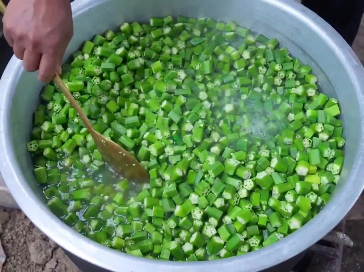 Des gombos verts coupés en train dêtre blanchis dans de leau bouillante à lintérieur dune grande casserole tout en étant remués avec une cuillère en bois.