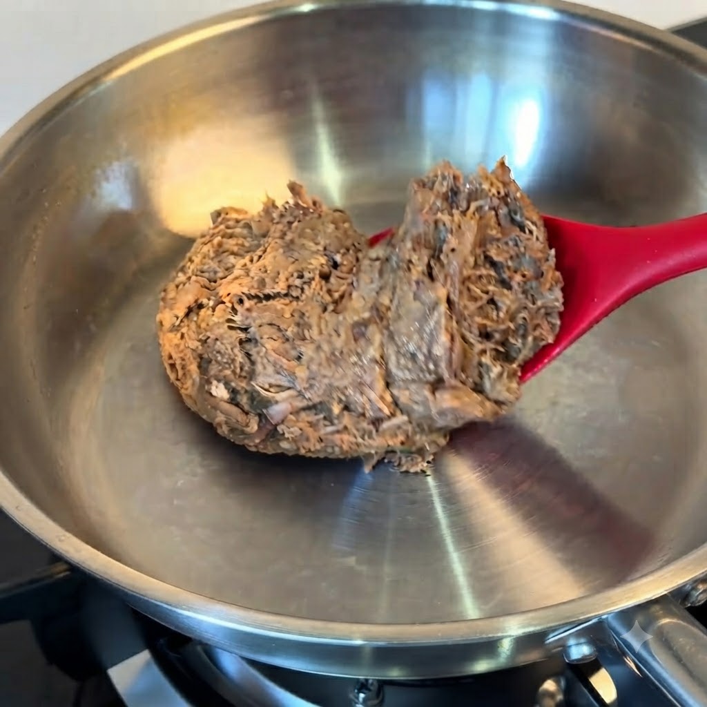 Un bloc de bœuf effiloché décongelé placé dans une poêle en acier inoxydable à laide dune spatule rouge.