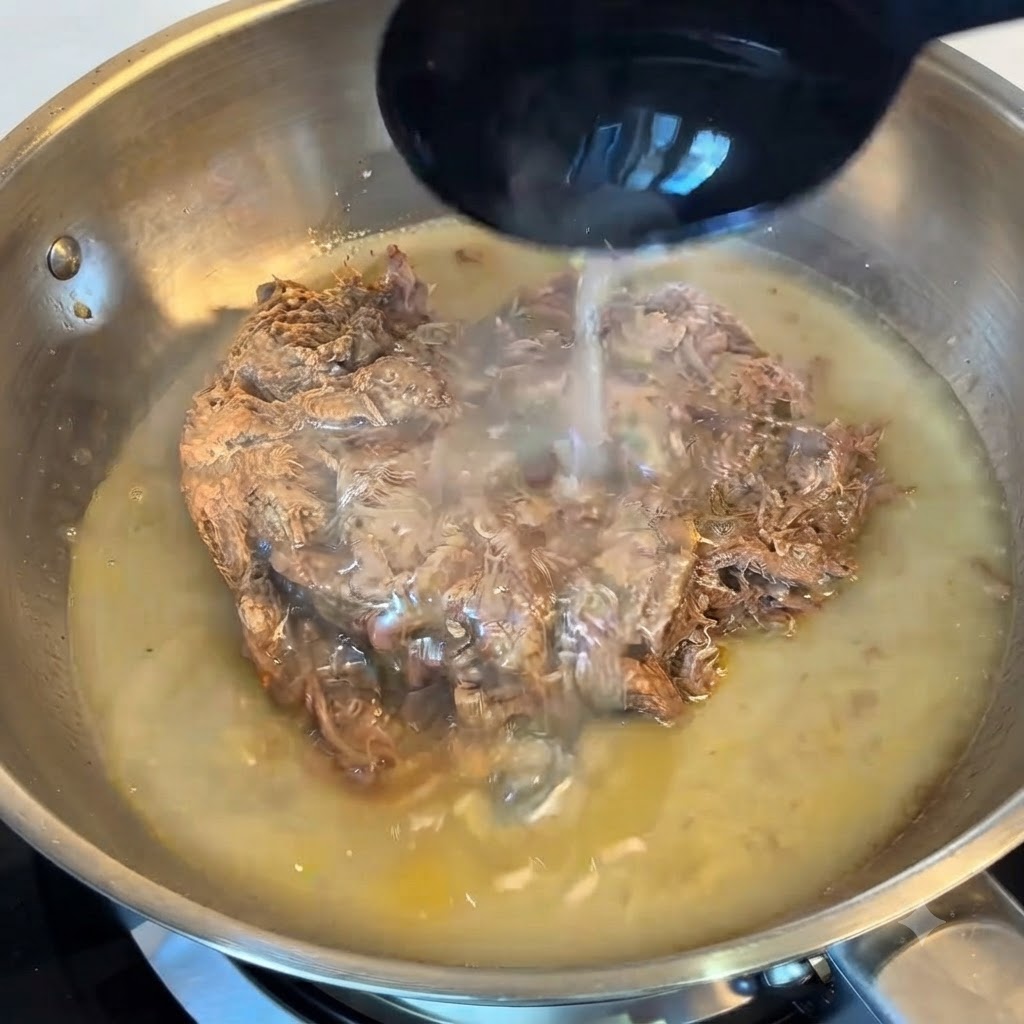 Une louche versant de leau de cuisson amidonnée sur le bœuf effiloché dans une poêle en acier inoxydable.