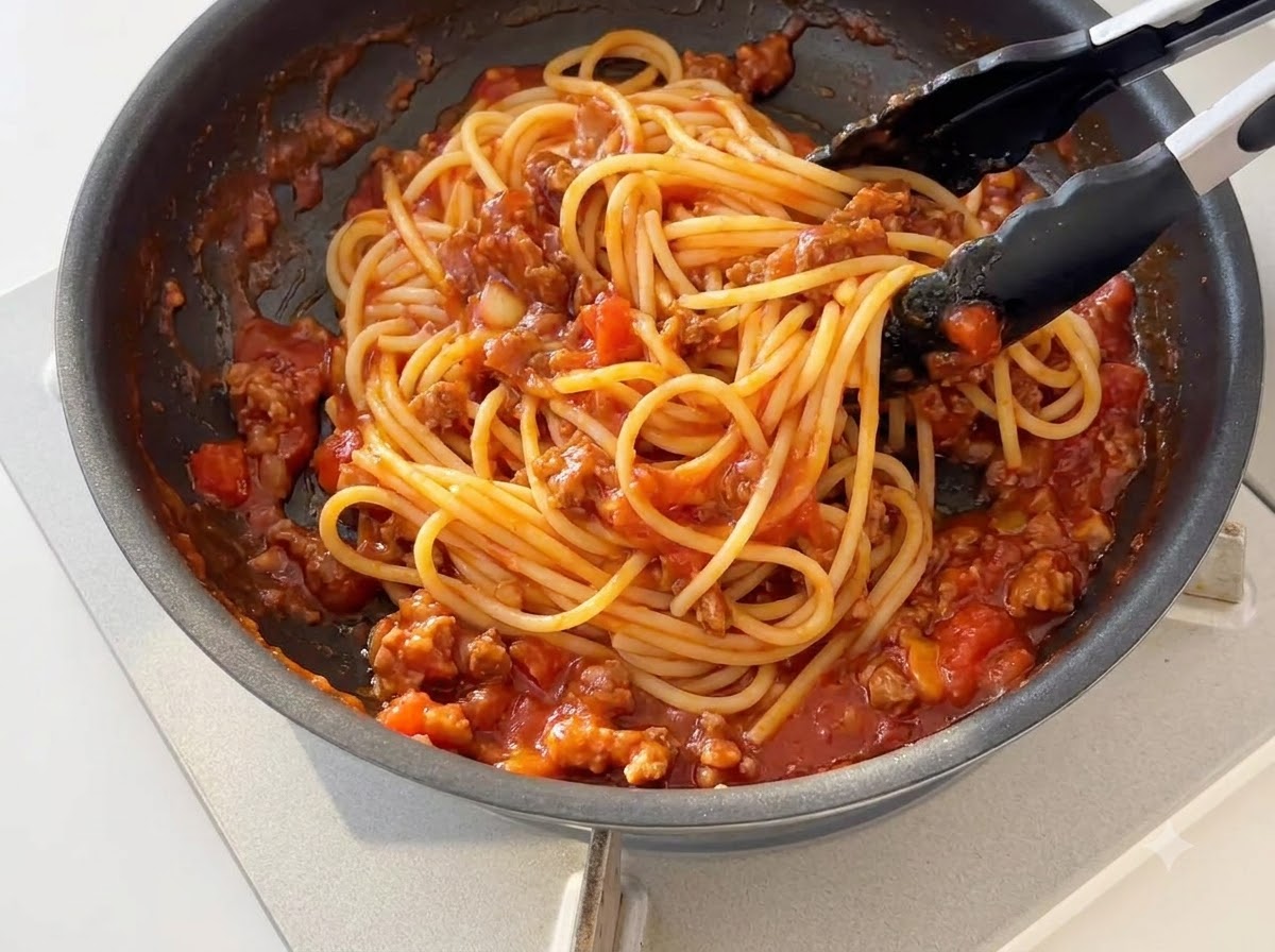 Des nouilles spaghetti cuites mélangées à la sauce tomate à la viande épaissie dans une poêle à laide de pinces en silicone noir.