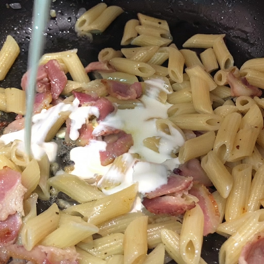 Du lait et de la crème entière versés par le haut dans une poêle remplie de penne et de morceaux de bacon.