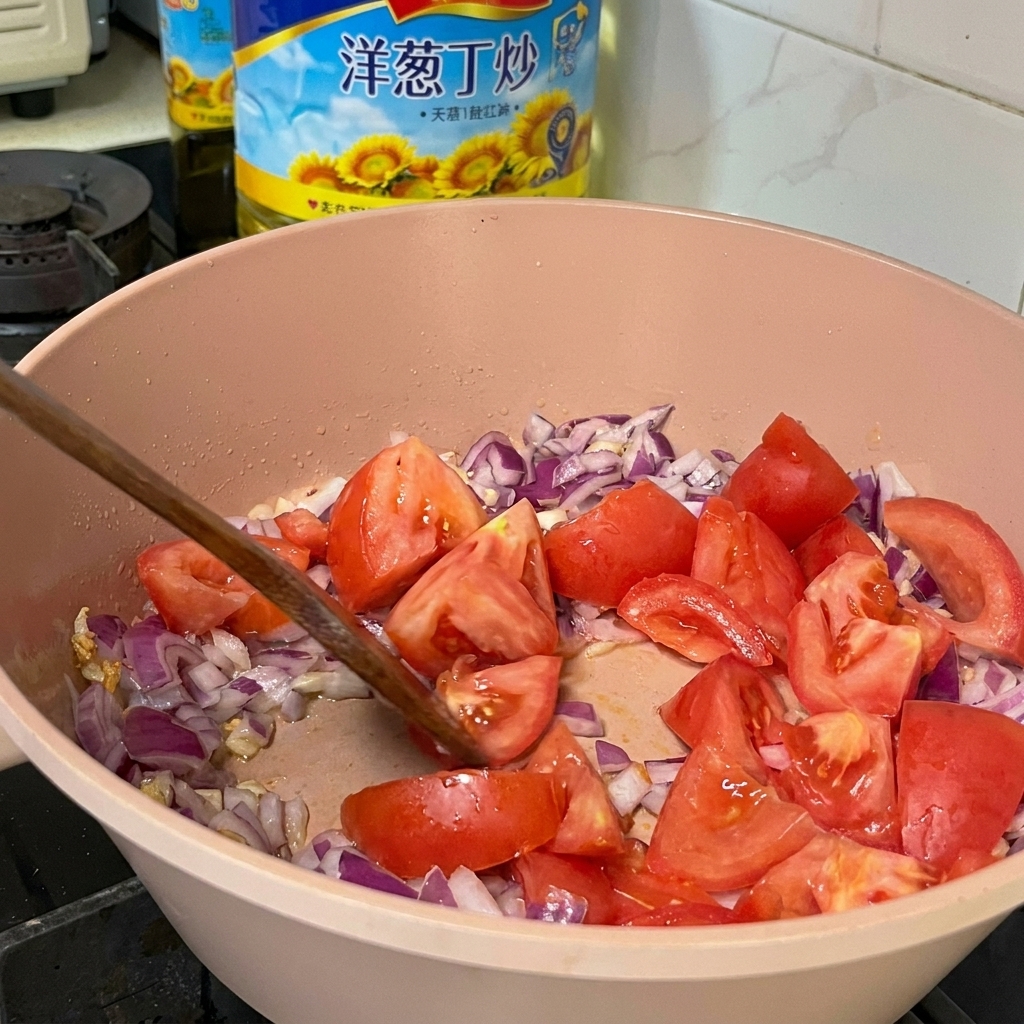 Morceaux de tomates fraîches mélangés à des oignons rouges en dés et de lail dans une poêle rose à laide dune spatule en bois.