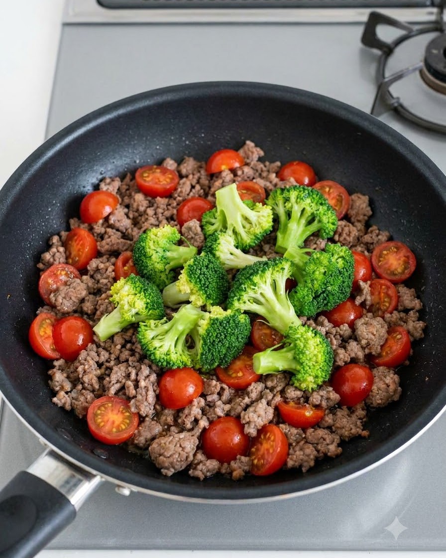 Des bouquets de brocoli vert et des tomates cerises rouge vif éparpillés sur le bœuf haché cuit dans une poêle.