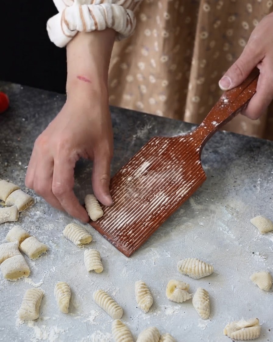 Main roulant un petit morceau de pâte sur une planchette à gnocchi en bois rainurée.
