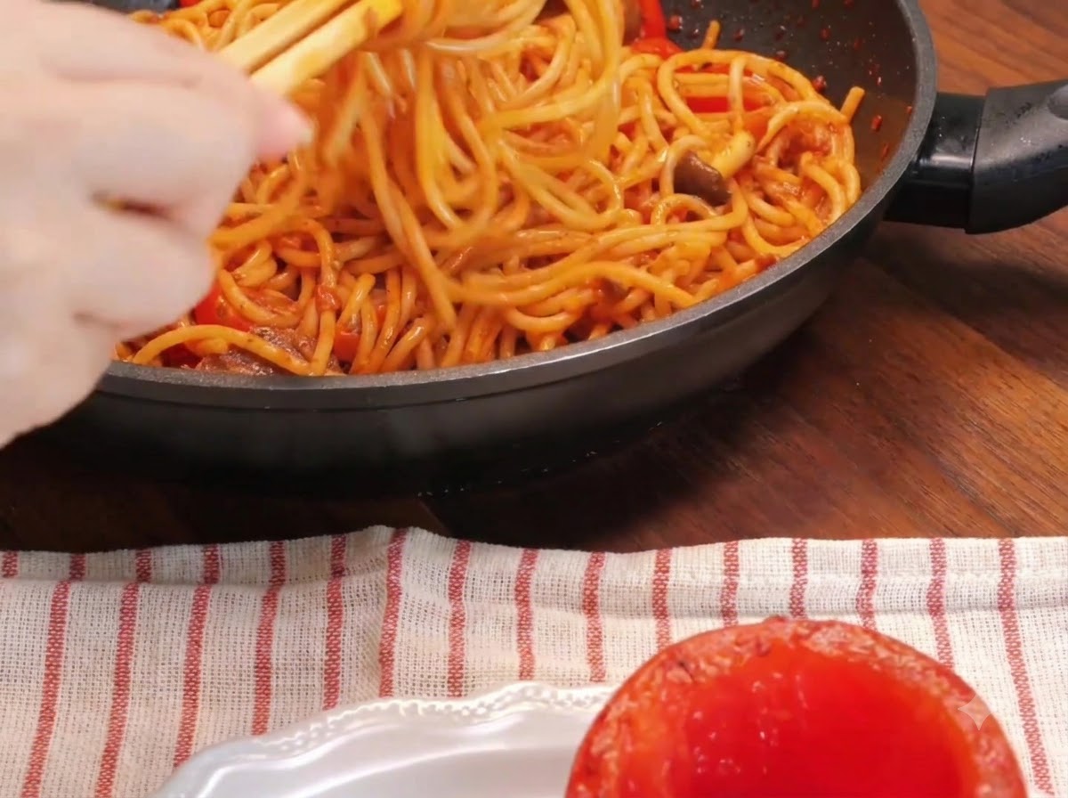 Gros plan dune main utilisant des baguettes pour tordre des nouilles spaghetti à lintérieur dune poêle à côté dune tomate rôtie évidée sur une assiette.