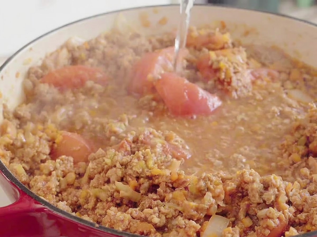 De leau versée du dessus dans une grande cocotte rouge contenant le mélange de viande, de tomates et de légumes en train de mijoter.