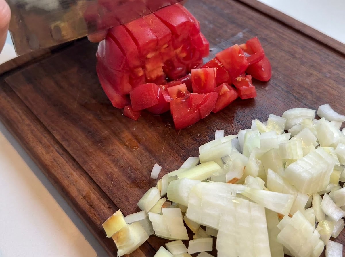 Une tomate rouge fraîche coupée en dés avec un couteau de cuisine sur une planche à découper en bois à côté doignons hachés.