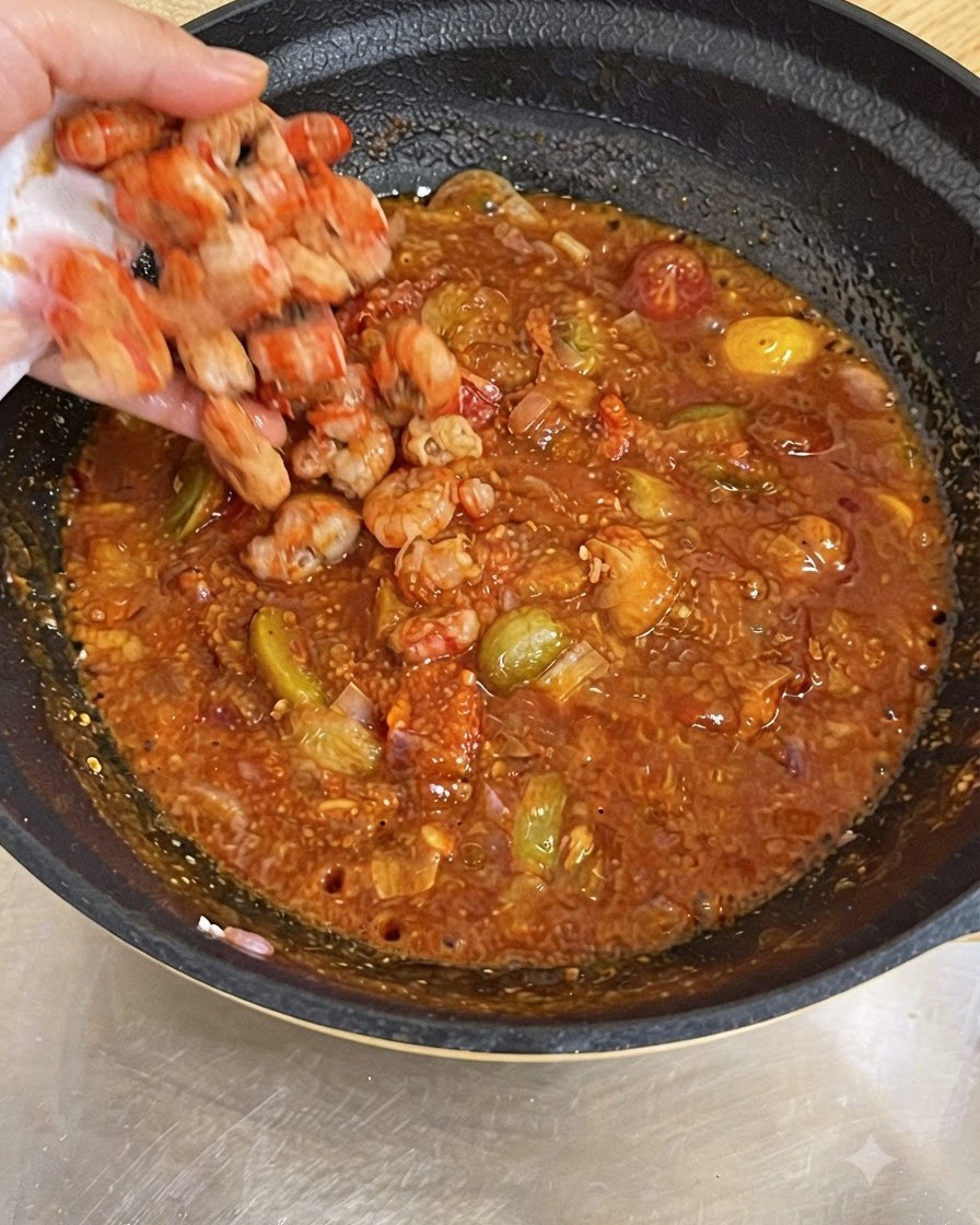 Une main jetant des crevettes cuites dans une poêle remplie de sauce tomate rouge bouillonnante.