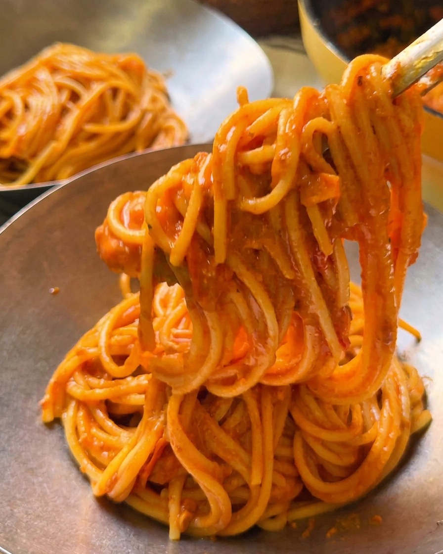 Des pinces soulevant une portion de spaghettis à la tomate crémeuse dans un bol de service en métal poli.
