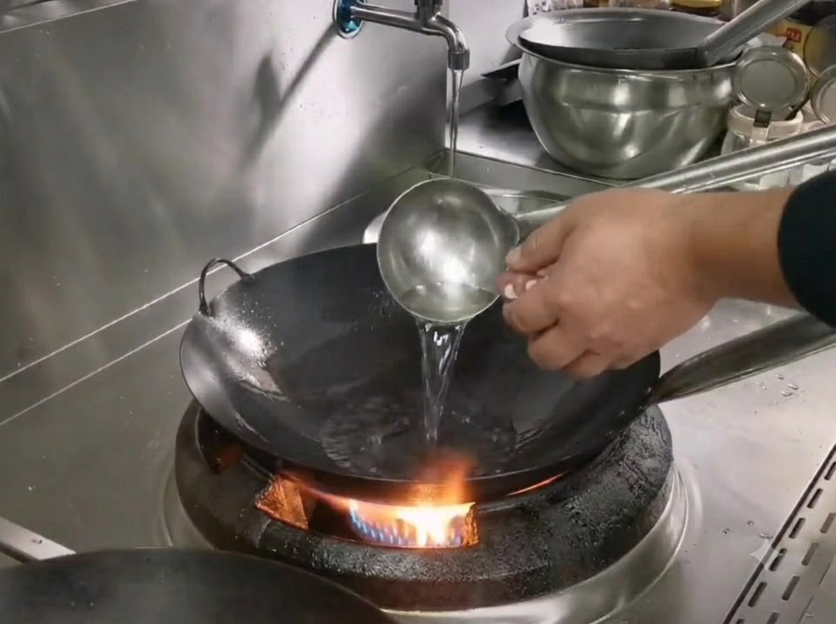 Une louche versant de lhuile dans un wok en fer chaud sur un brûleur à gaz tout en préparant la cuisson du porc émincé.