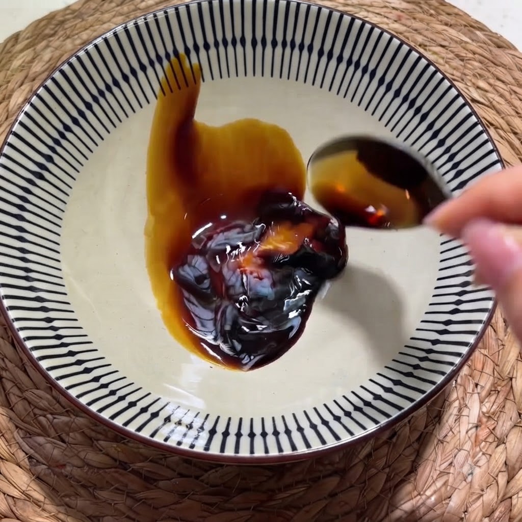 Une main versant de la sauce soja foncée à partir dune cuillère dans un bol en céramique à motifs contenant une cuillerée de sauce huître.