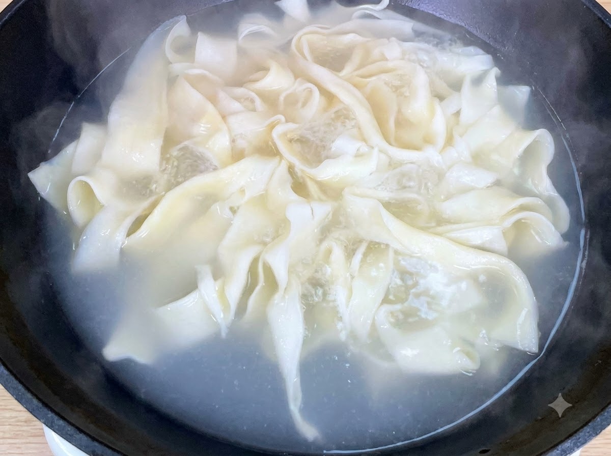 Du bok choy frais bouillant aux côtés de nouilles larges dans une casserole.