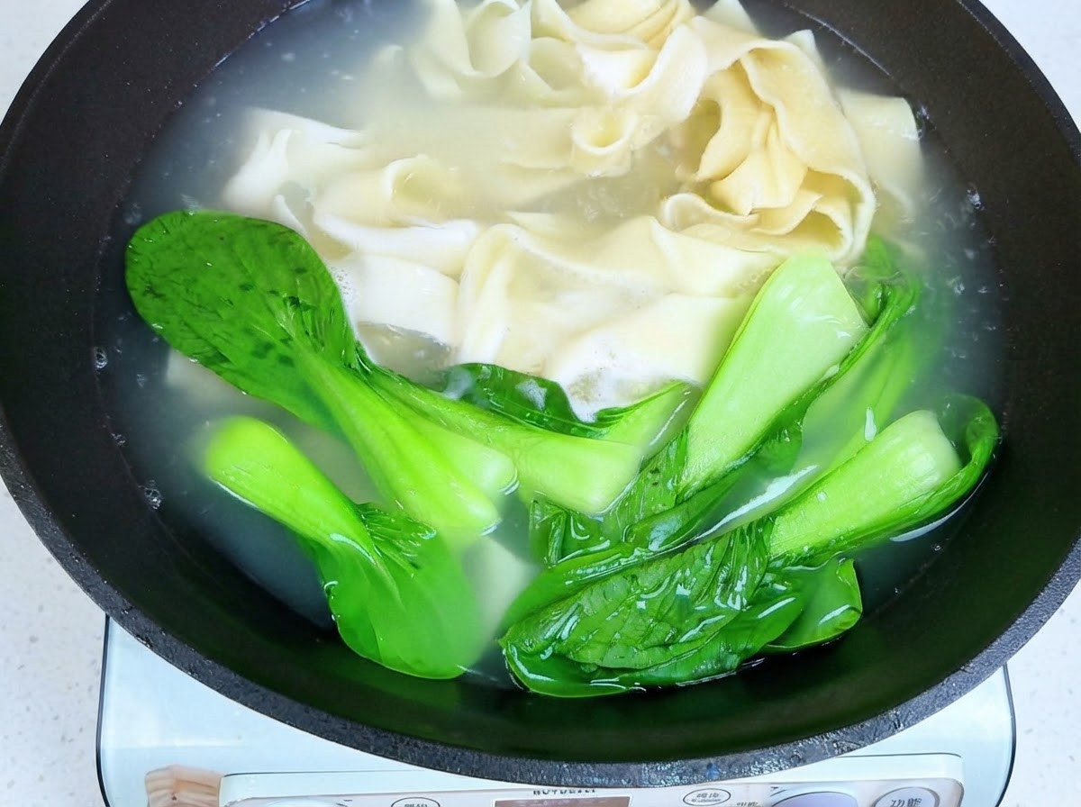 Des baguettes soulevant des nouilles larges cuites et du bok choy hors de leau chaude.