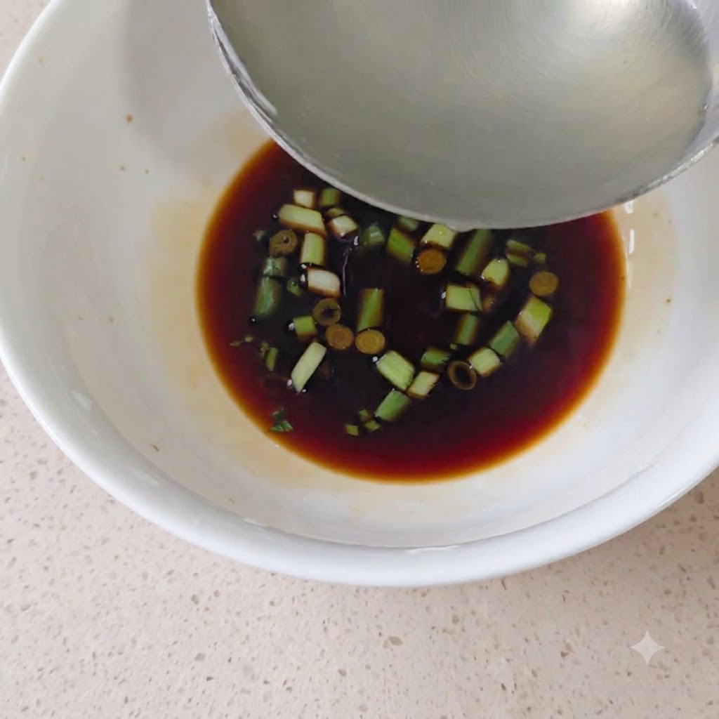 Une louche en métal versant du bouillon chaud dans le bol rempli de sauce soja et de ciboule.